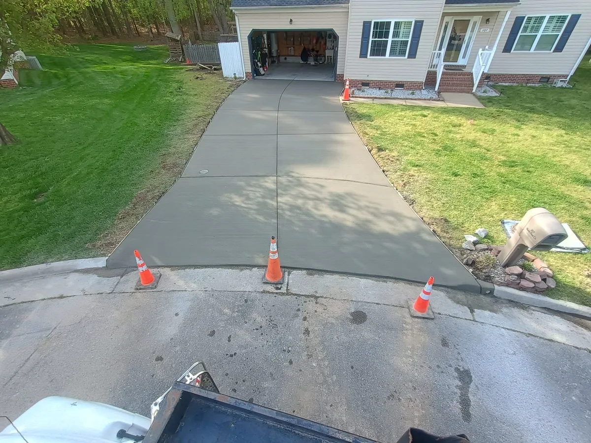 Newly poured concrete driveway with control joints leading to a suburban home's garage, bordered by green lawns and marked with orange traffic cones at the curb