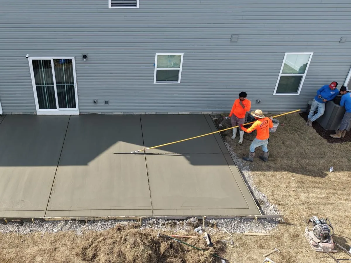 Aerial view of construction workers smoothing a freshly poured concrete patio in a backyard beside a gray house