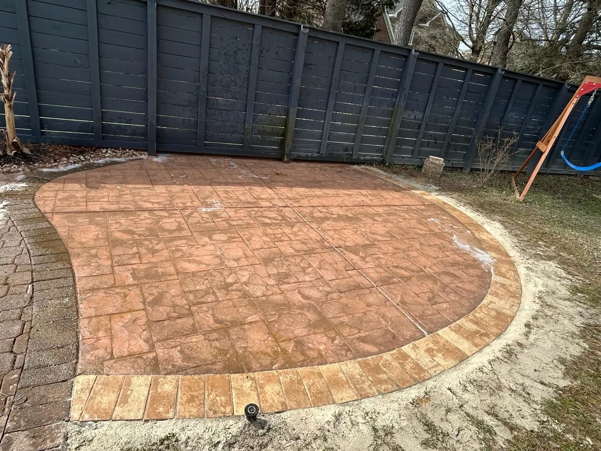 A stamped concrete patio with a decorative border sits in a backyard beside a dark wooden fence