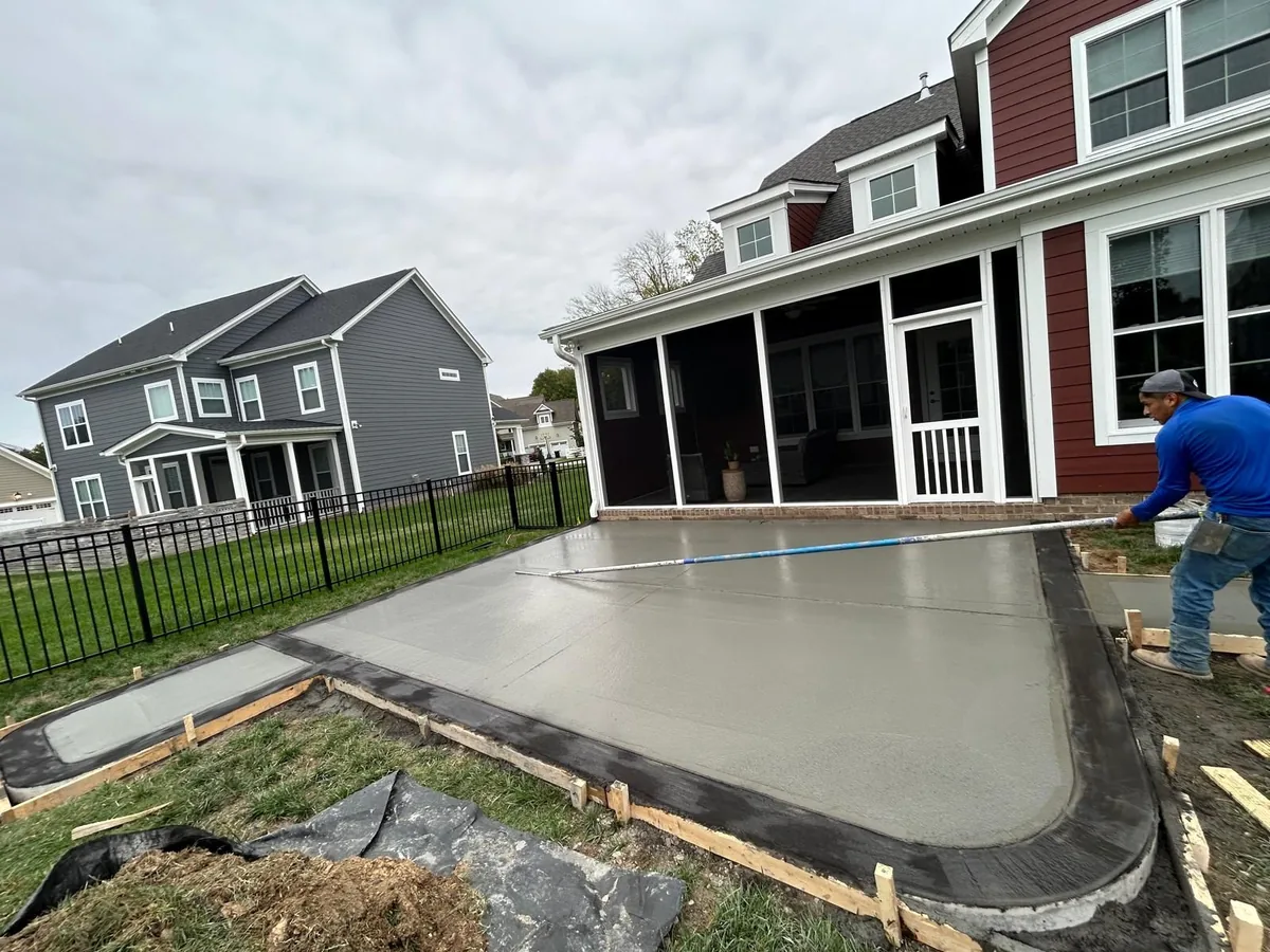 Worker smoothing a newly poured concrete patio in a backyard next to a house with a screened porch